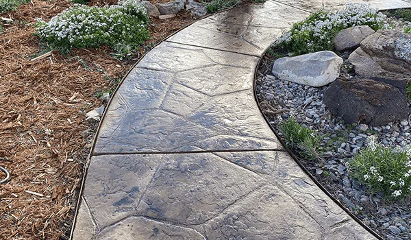 Stamped concrete sidewalk with natural stone pattern surrounded by plants and gravel, showcasing the design versatility of concrete compared to pavers