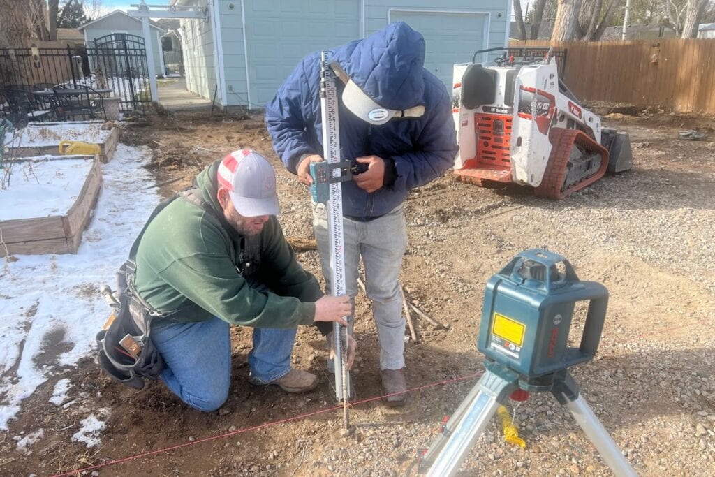 Contractors performing excavation and laser level grading for reinforced 6-inch concrete driveway replacement and extension in Windsor, CO, preparing compacted base for heavy-duty 4500 PSI slab installation.