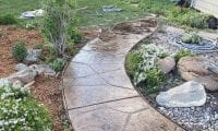 A curved concrete walkway with a stamped stone pattern winds through a front yard. The walkway is bordered by rocks, and there are flowering plants and shrubs nearby.