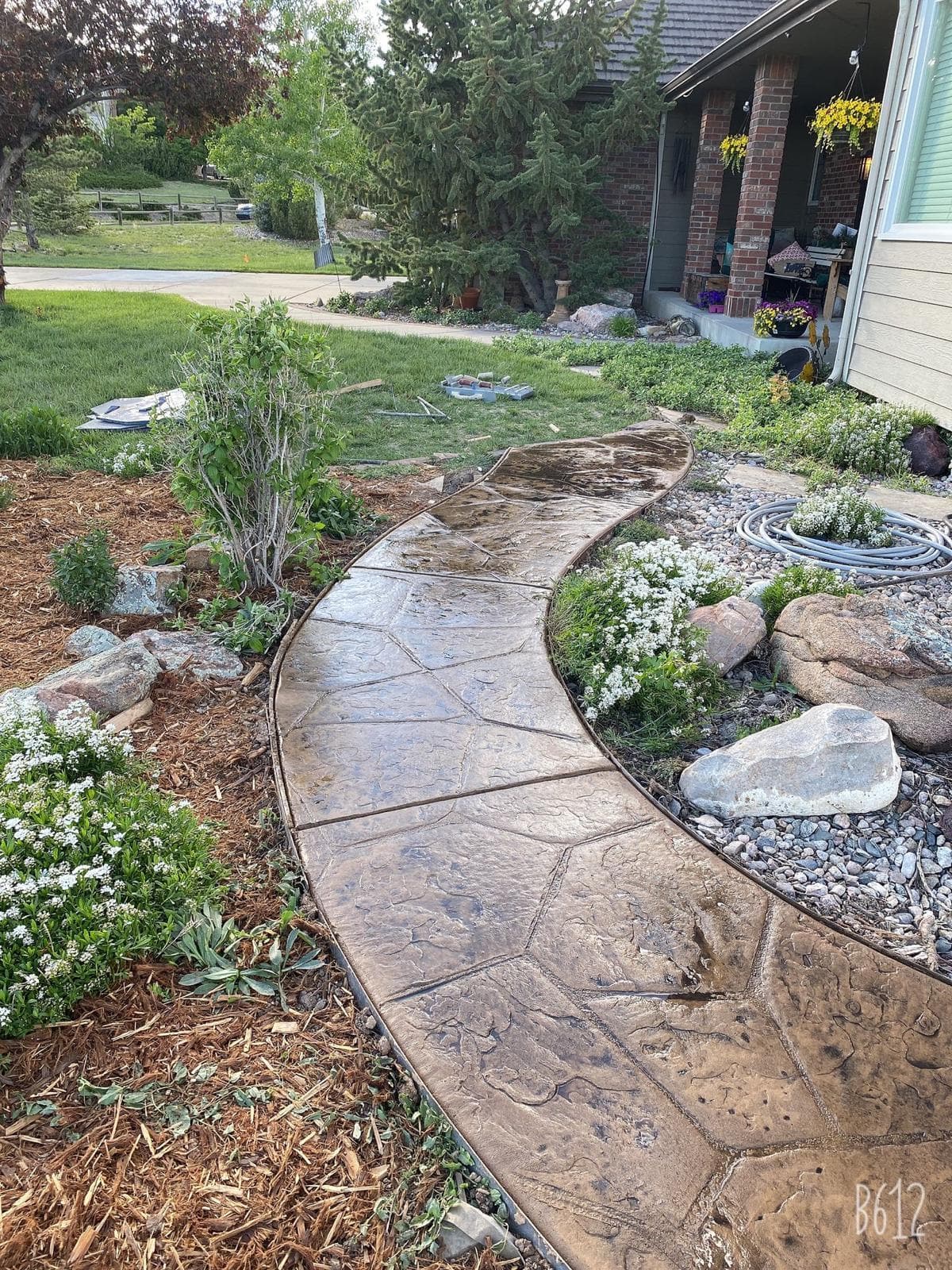 A curved concrete walkway with a stamped stone pattern winds through a front yard. The walkway is bordered by rocks, and there are flowering plants and shrubs nearby.