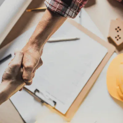 Concrete contractor shaking hands with a client over project plans, with a hard hat and construction documents on the table.