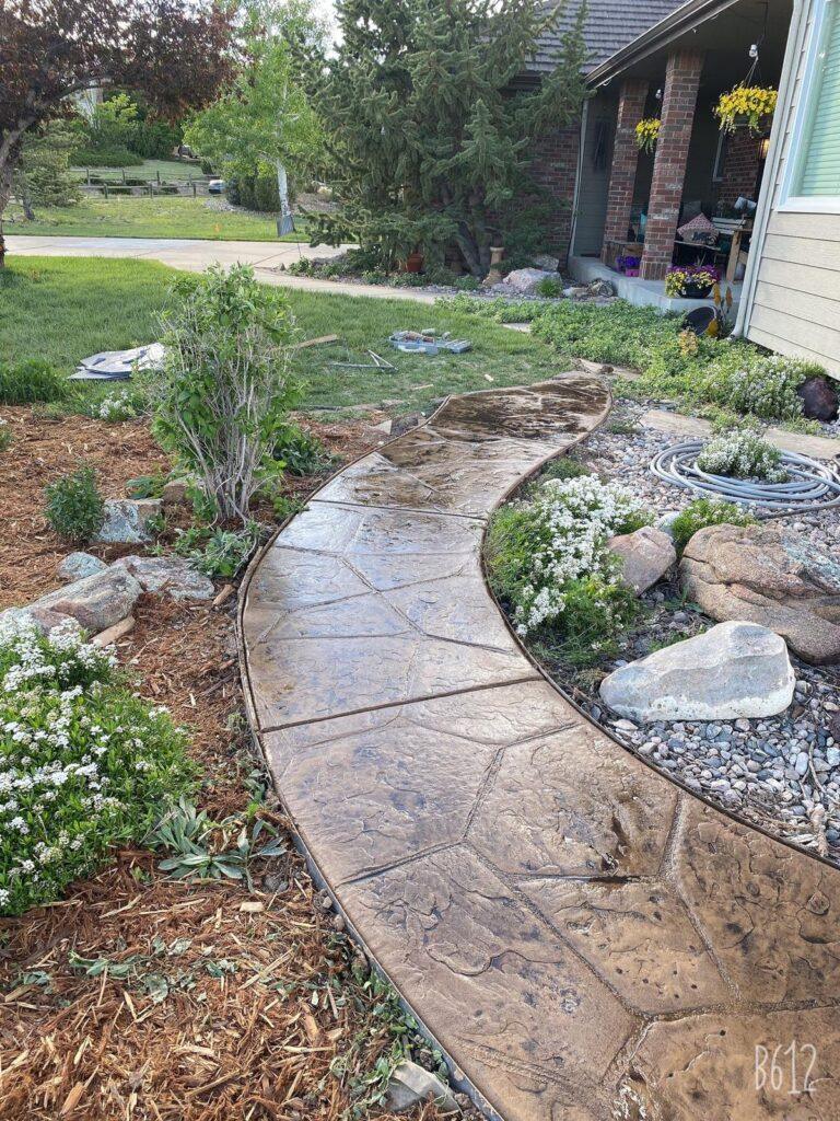 A curved concrete walkway with a stamped stone pattern winds through a front yard. The walkway is bordered by rocks, and there are flowering plants and shrubs nearby.