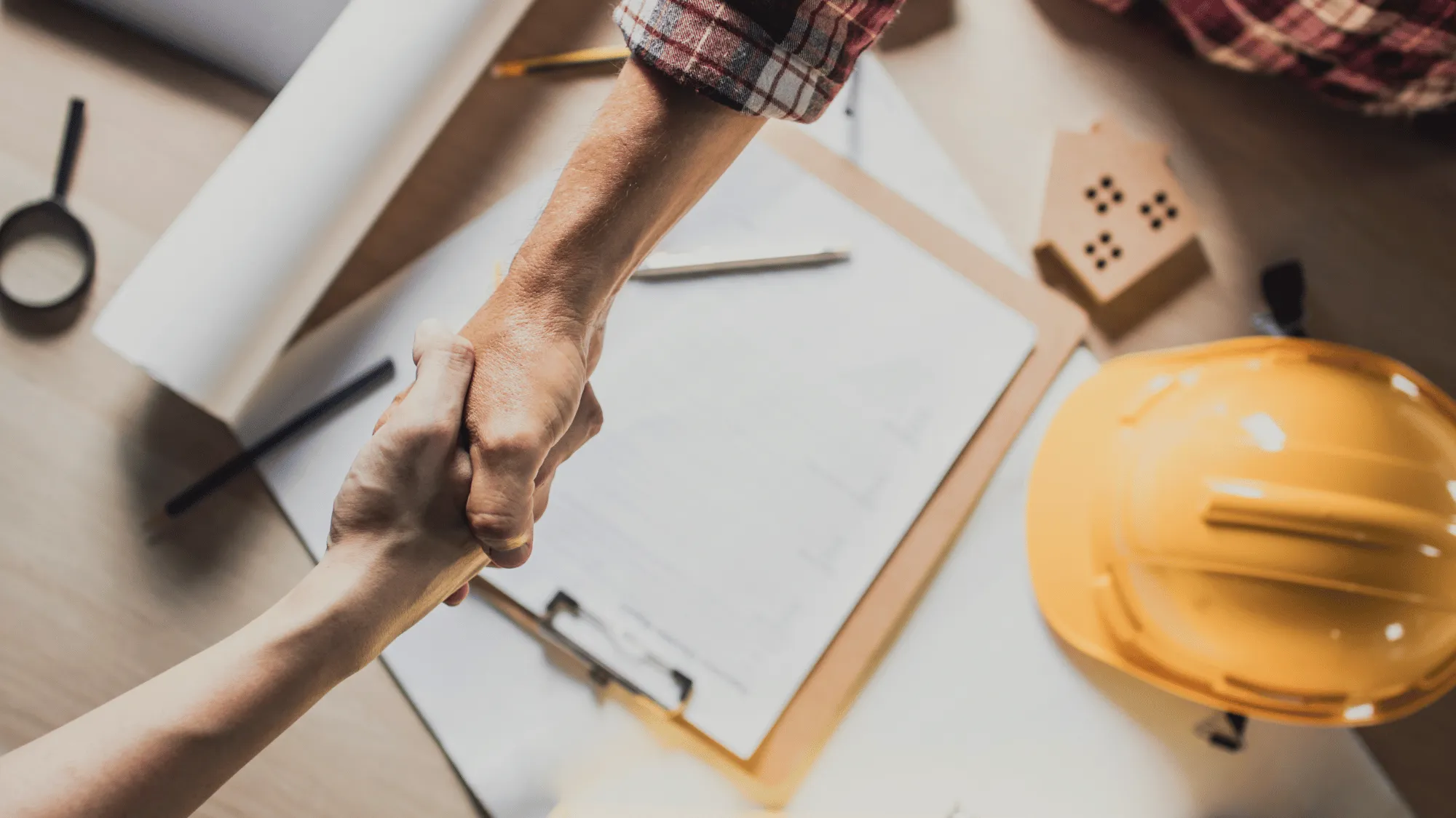 Concrete contractor shaking hands with a client over project plans, with a hard hat and construction documents on the table.