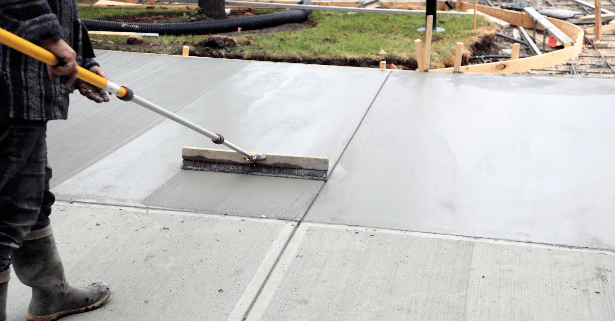 Worker smoothing freshly poured concrete with a finishing broom on a residential sidewalk or patio project.